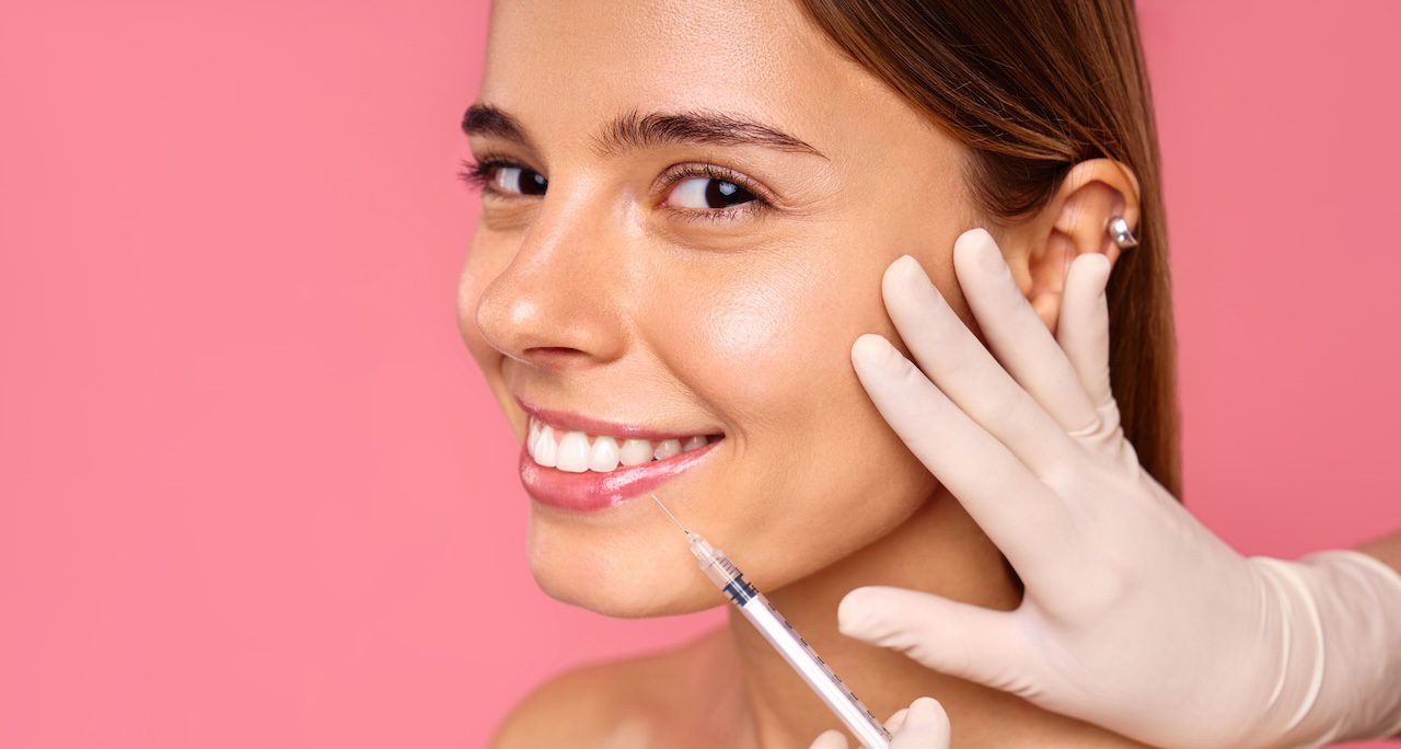 smiling woman receiving a cosmetic injection in her cheek against a pink background