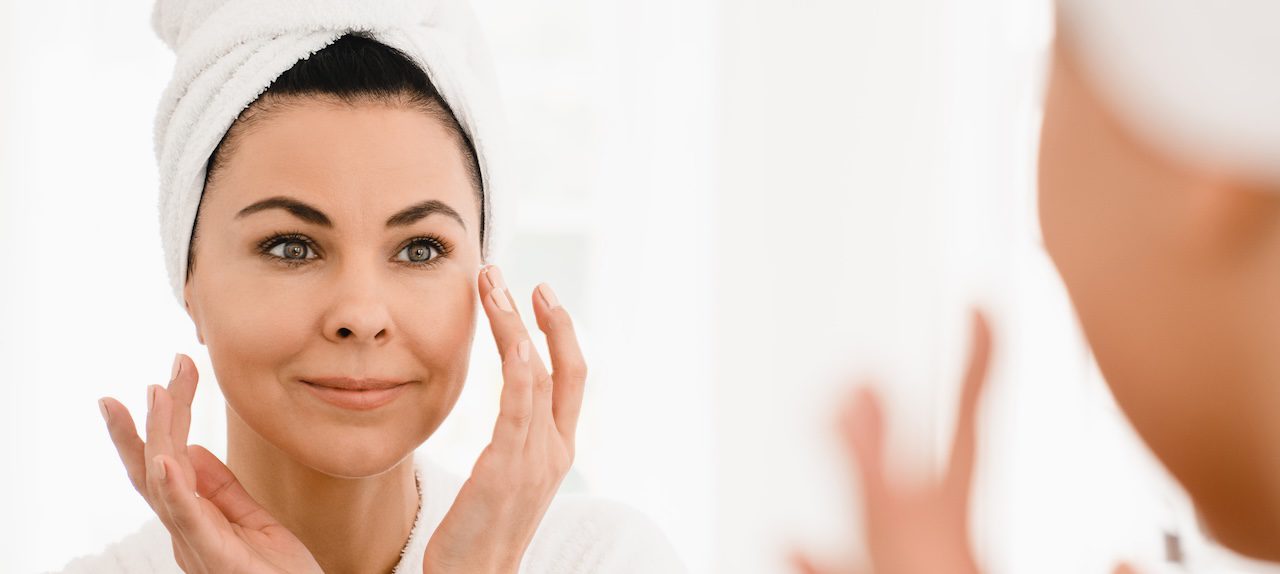 woman in a robe and hair towel touching her cheeks in front of a mirror